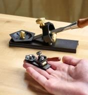 The miniature honing guide is held on the fingers of an open hand while the full-size honing guide appears in the background