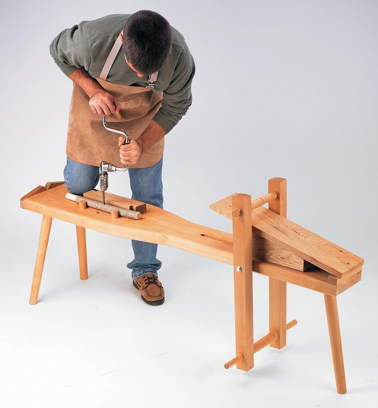 A man kneeling on a shaving horse uses a hand drill to bore a hole in a workpiece